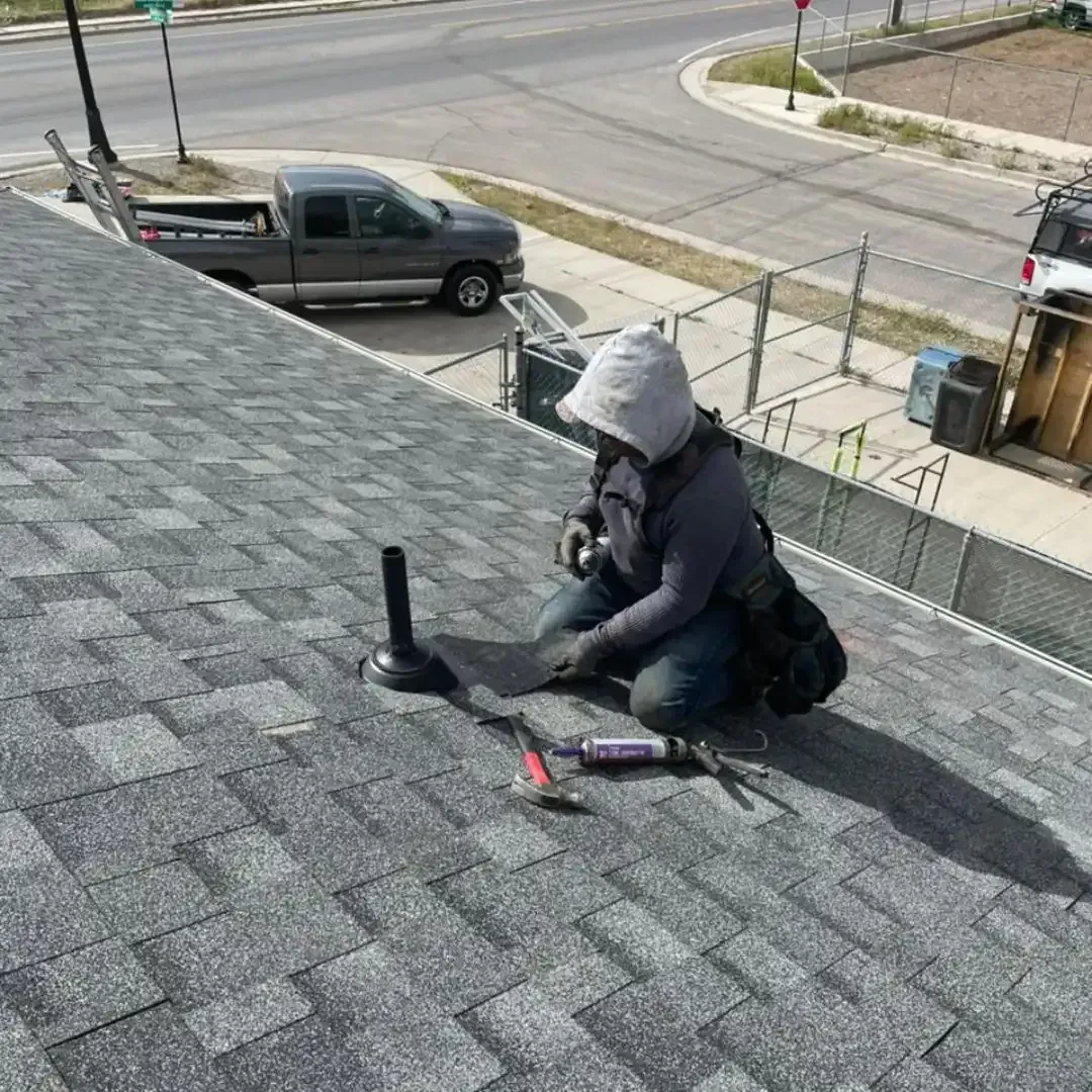 Roofer in hooded jacket kneeling on gray shingle roof, working on a vent. A truck is parked on the street below.