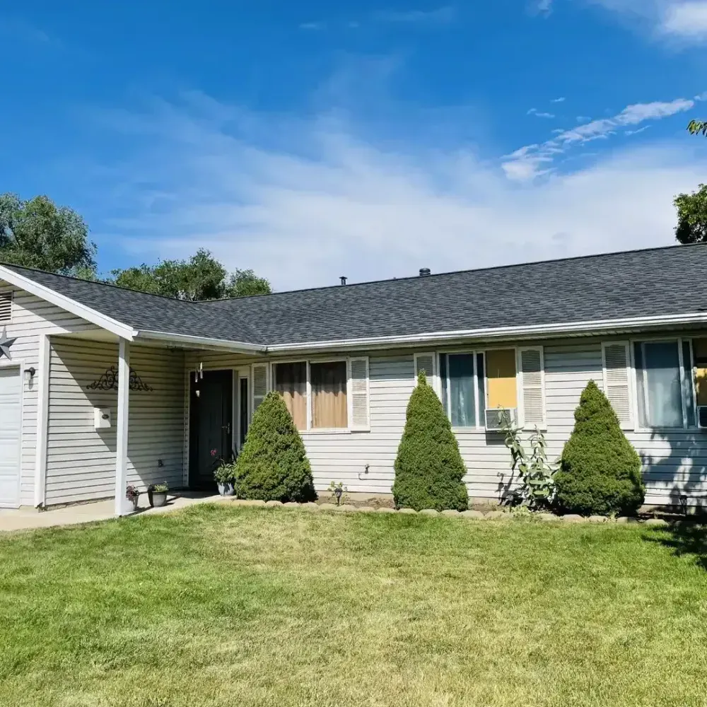 Ranch-style house with gray roof, beige siding, green lawn, three evergreen bushes, and blue sky.