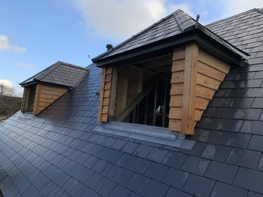 Wooden dormers under construction on a dark gray slate roof against a blue sky.