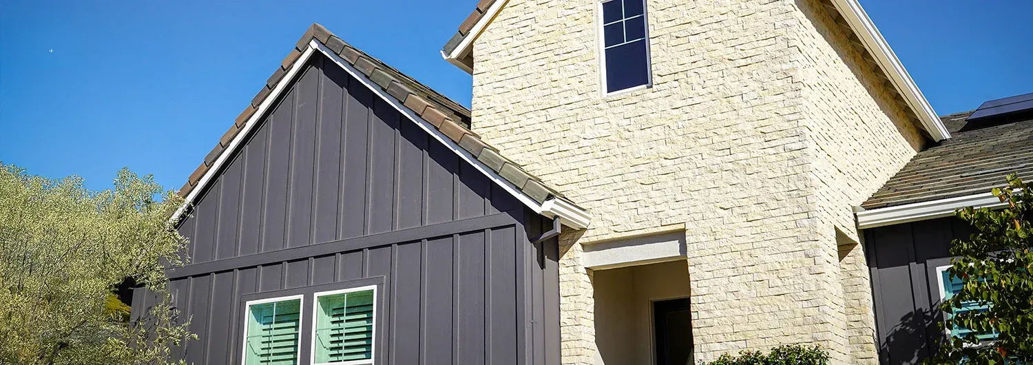 Modern home exterior with dark vertical board-and-batten siding and a light-colored stone facade under a bright blue sky.