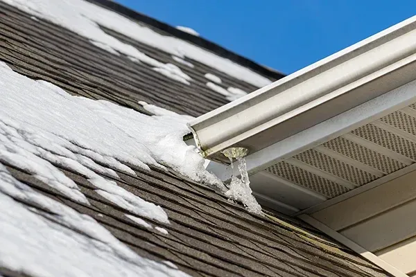 Snow-covered roof with ice stalactites hanging from the gutter in front of a blue sky.