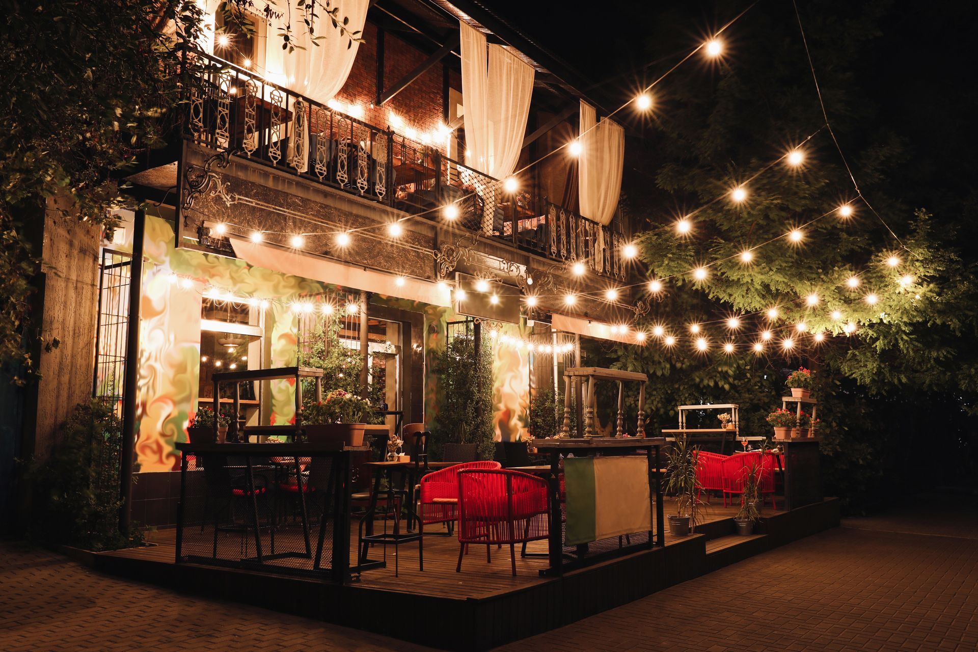 Nighttime outdoor restaurant with string lights, red chairs, and a balcony.