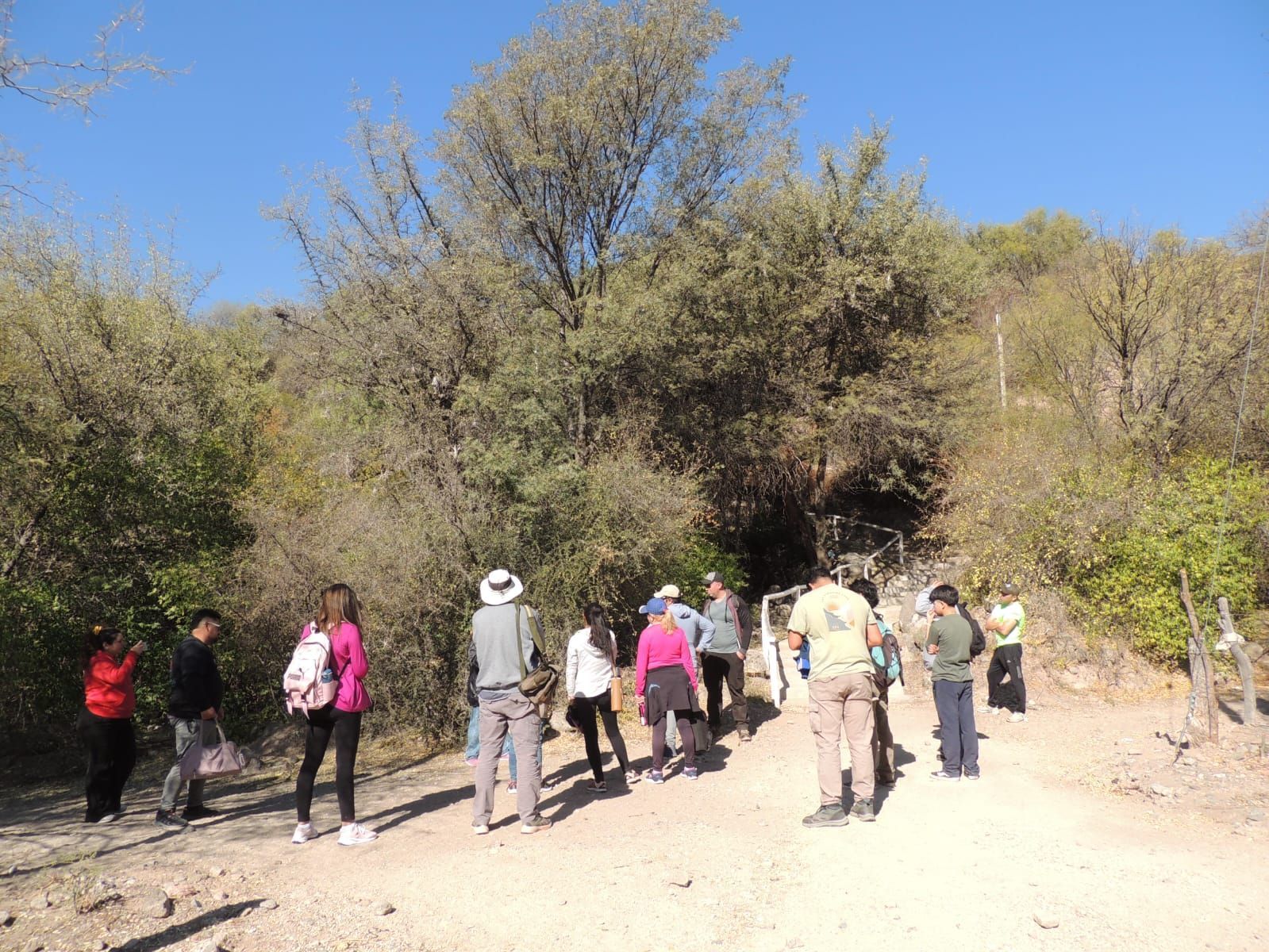 Grupo de personas caminando por un sendero de tierra, rodeado de árboles y arbustos, bajo un cielo azul.