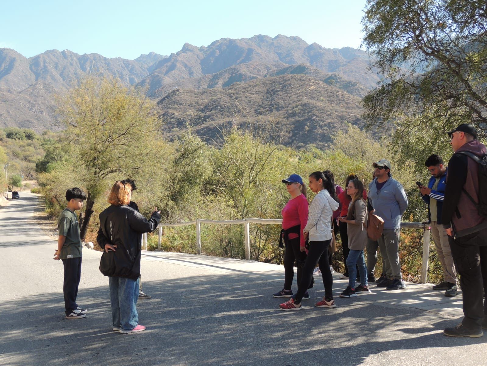 Grupo de personas en una carretera, montañas al fondo, posiblemente una visita guiada.