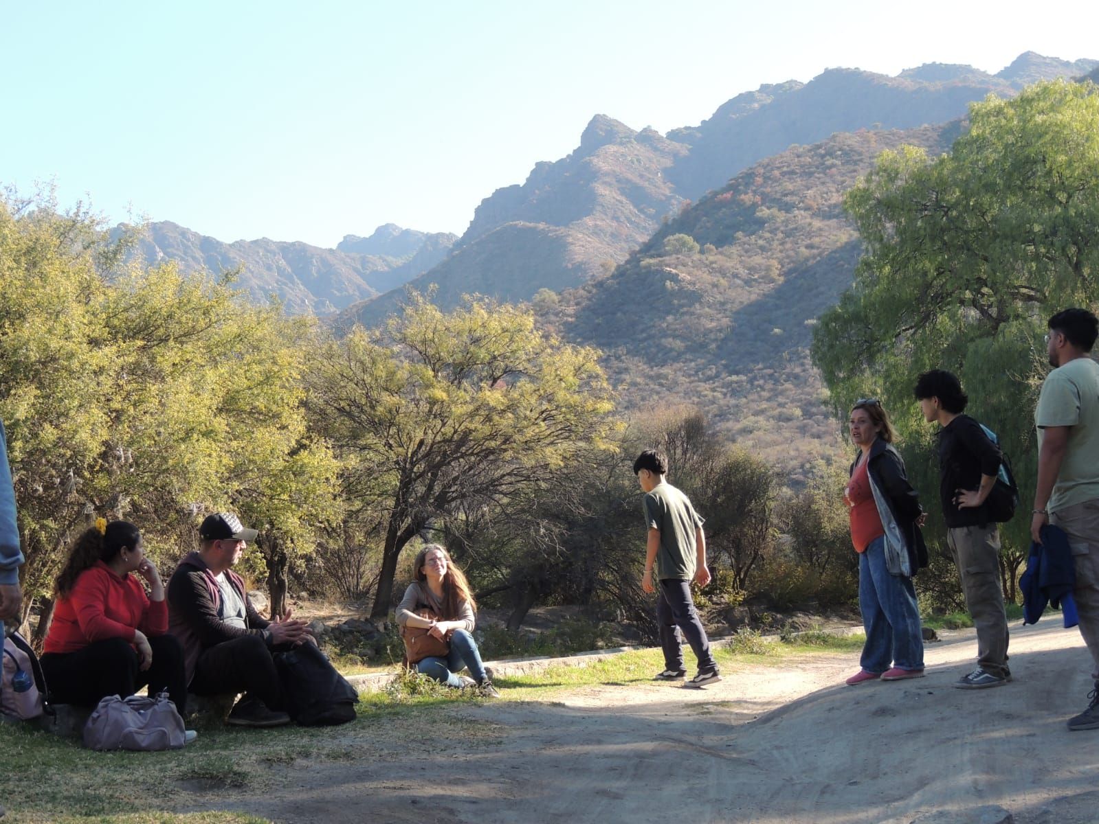 Grupo de personas al aire libre frente a las montañas; algunos de pie, otros sentados, interactuando.
