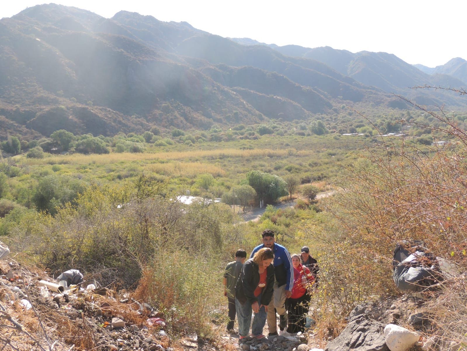 Personas caminando por un sendero hacia un valle con montañas al fondo. Un entorno soleado al aire libre.