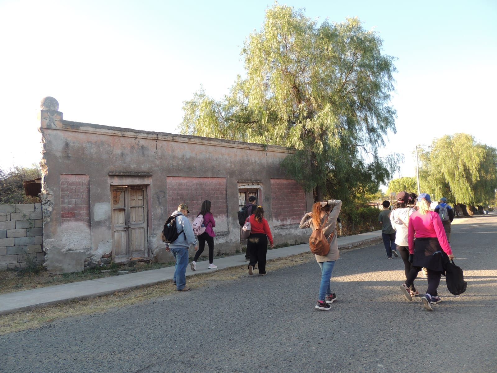 Gente caminando por la calle, mirando un edificio deteriorado. Árboles y cielo al fondo.