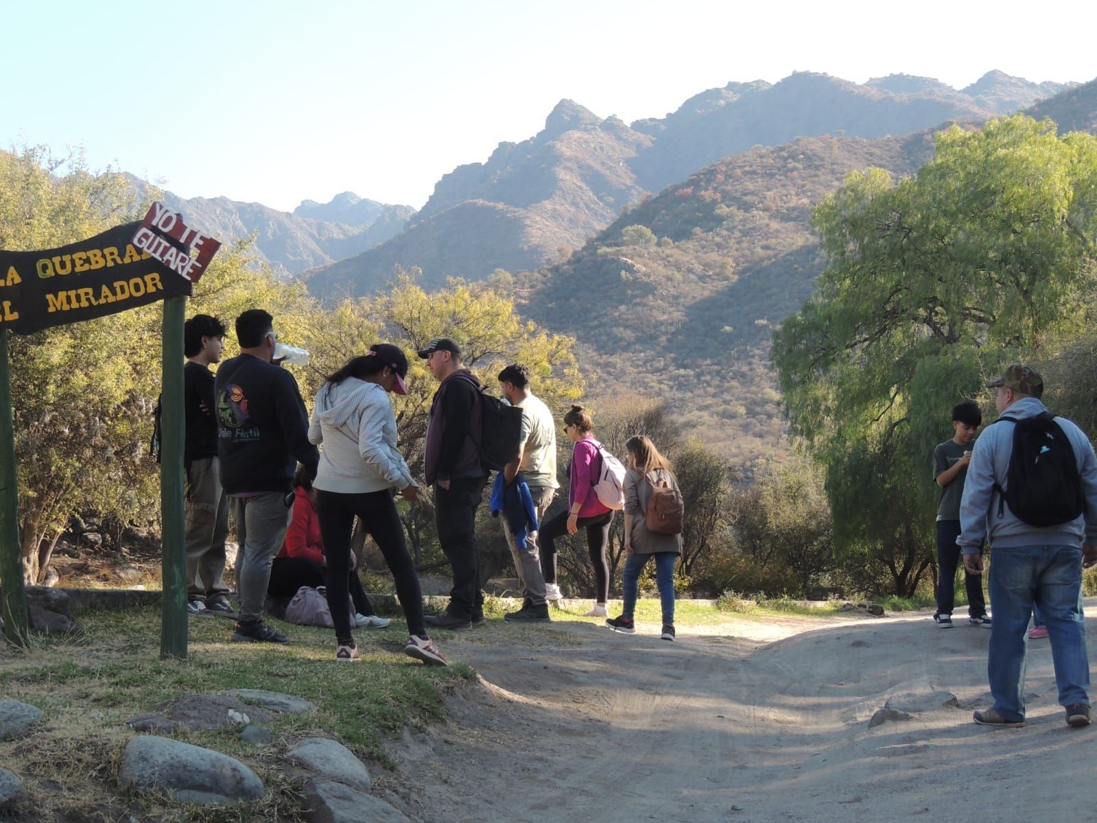Gente cerca de un sendero, con montañas al fondo. Luz del sol, árboles y un camino de tierra.