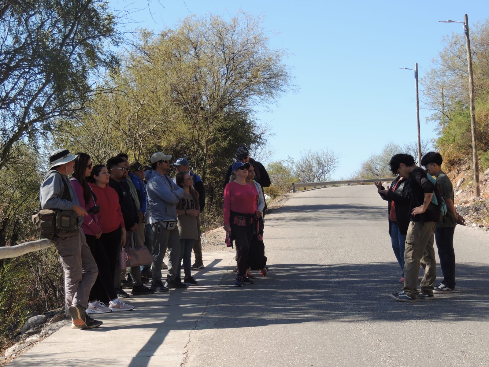 Gente parada en una calle pavimentada, posiblemente escuchando a un orador, árboles de fondo, día soleado.