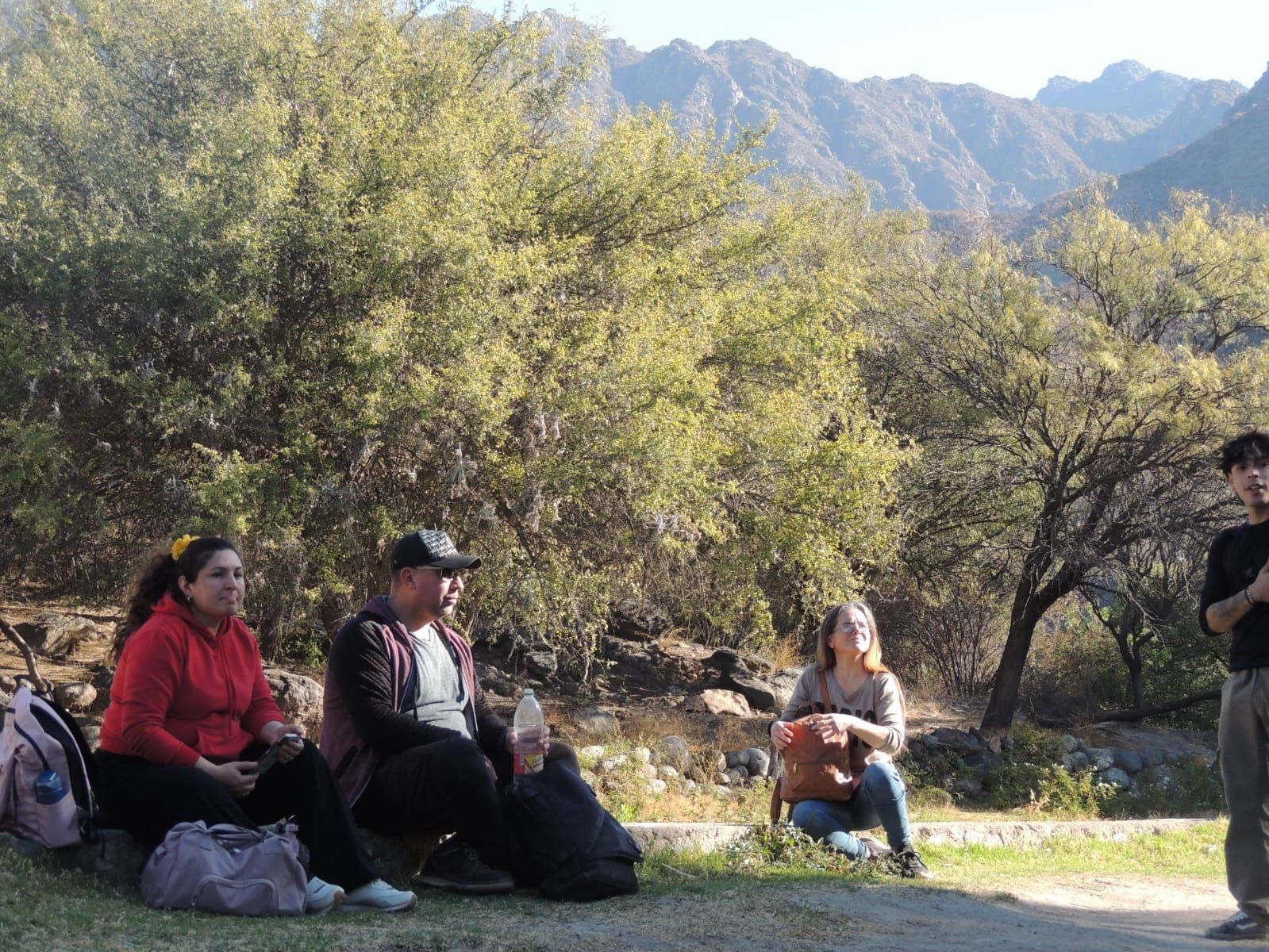 Personas sentadas y de pie cerca de árboles con un fondo de montaña.
