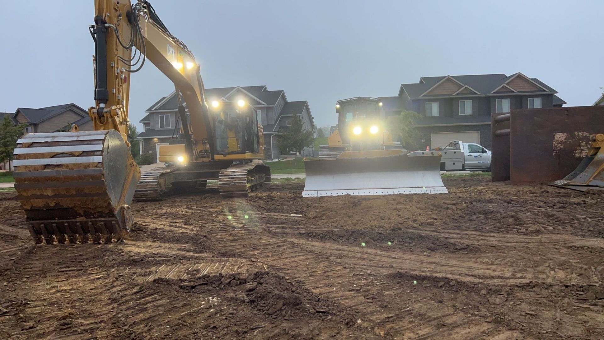 An excavator digging on a muddy construction site in front of houses at dusk.