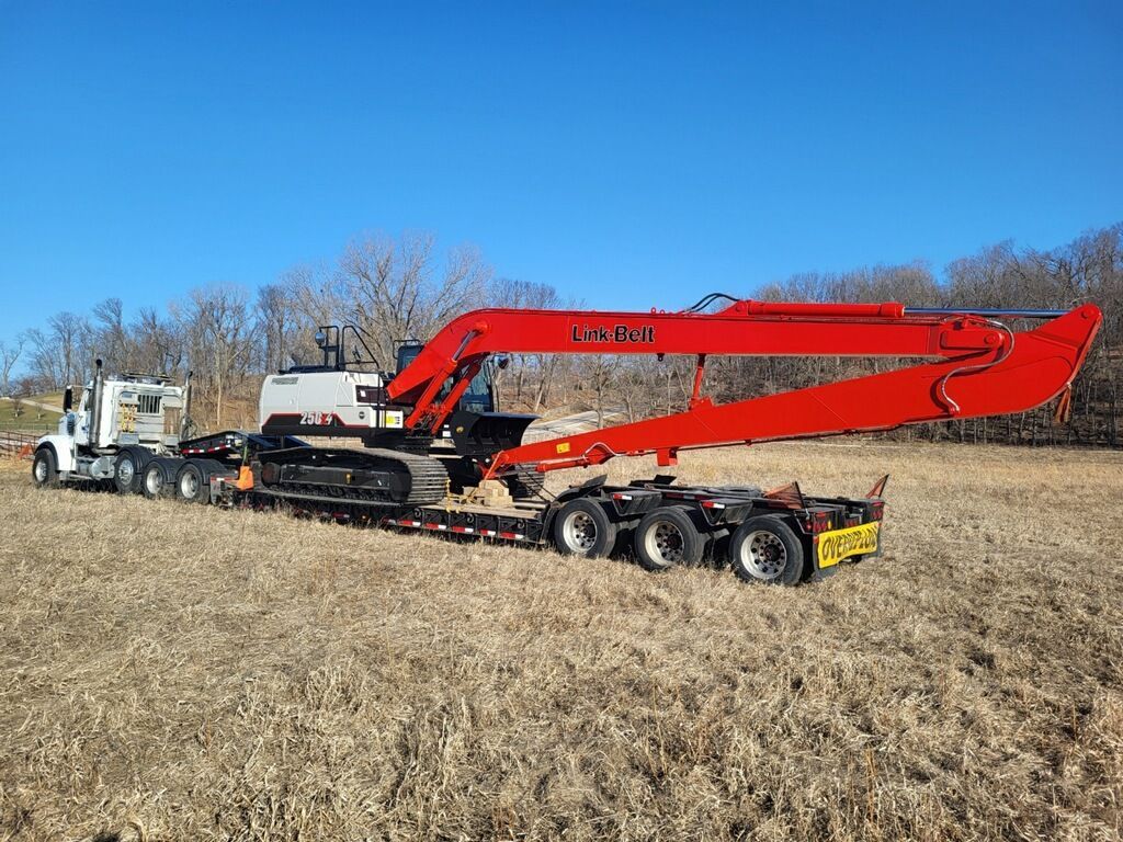 A red excavator on a trailer, pulled by a white truck, in a grassy field on a sunny day.