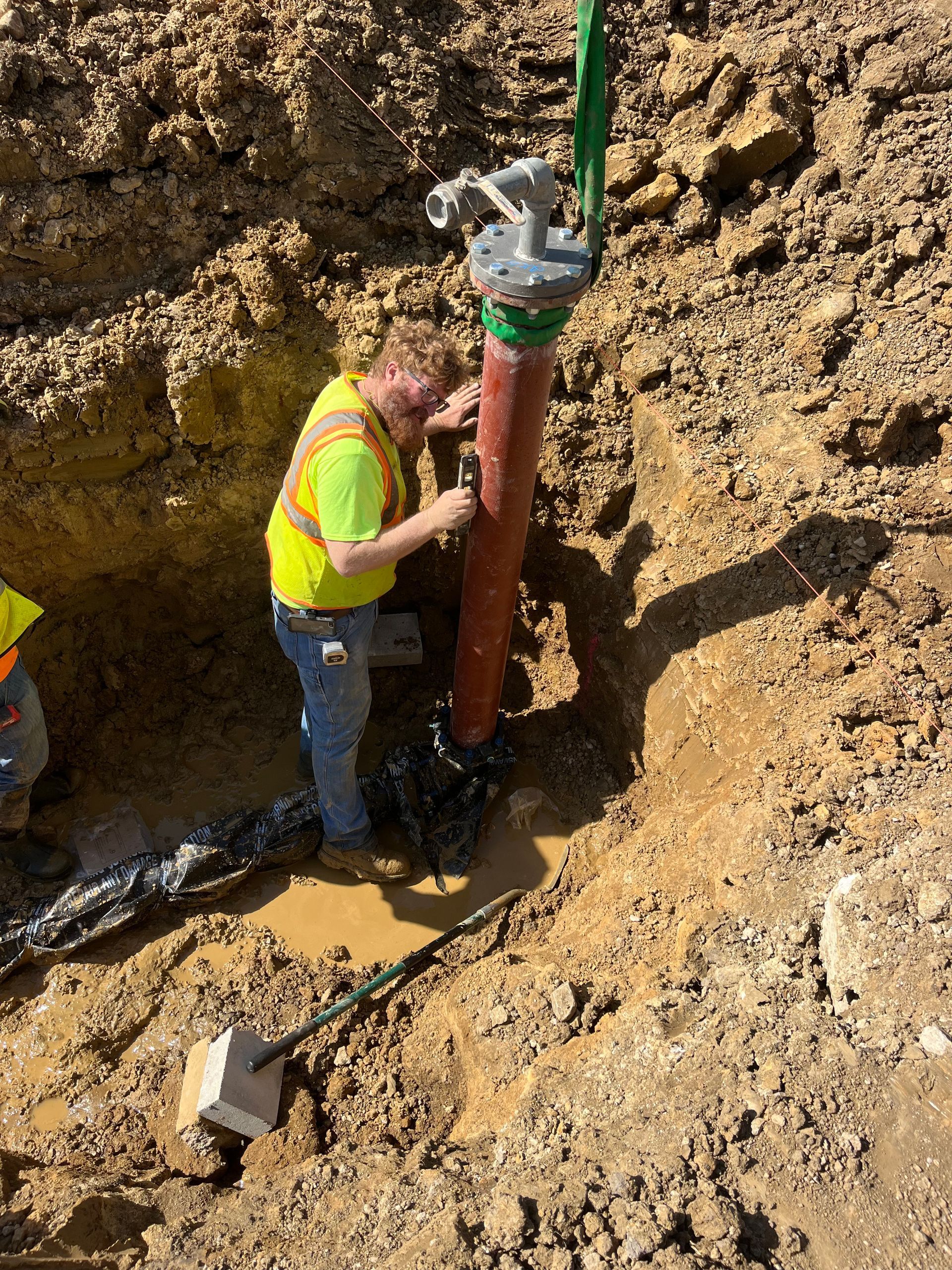 Man in a yellow vest repairing a pipe in a muddy trench. Earth and equipment are visible.
