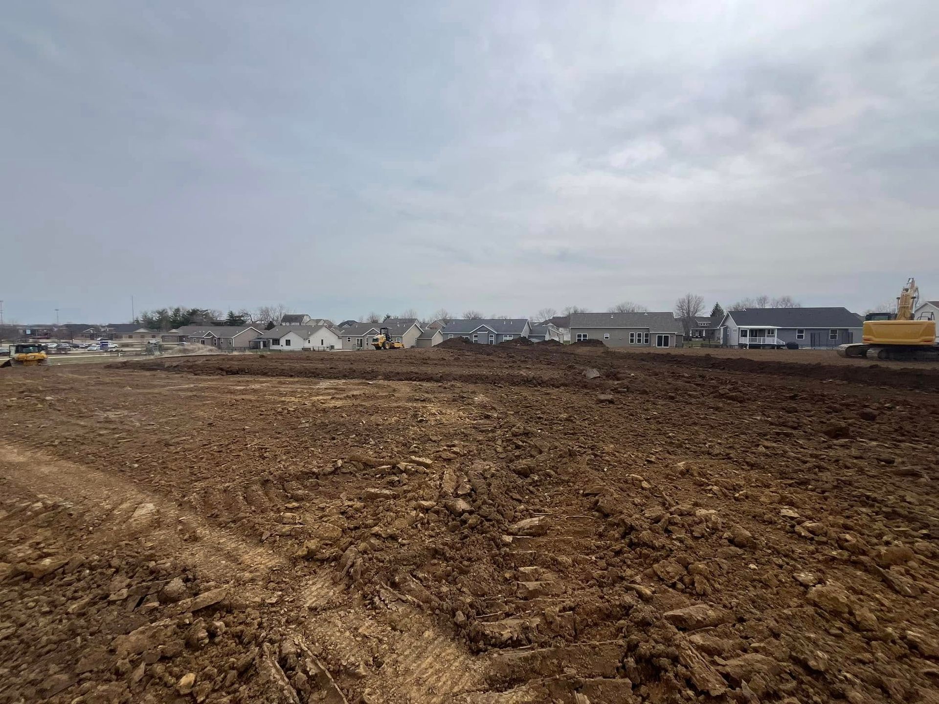 A large dirt field with houses in the background and a yellow excavator in the foreground.