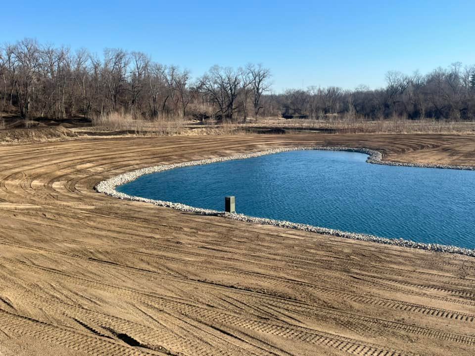A large pond in the middle of a dirt field with trees in the background.