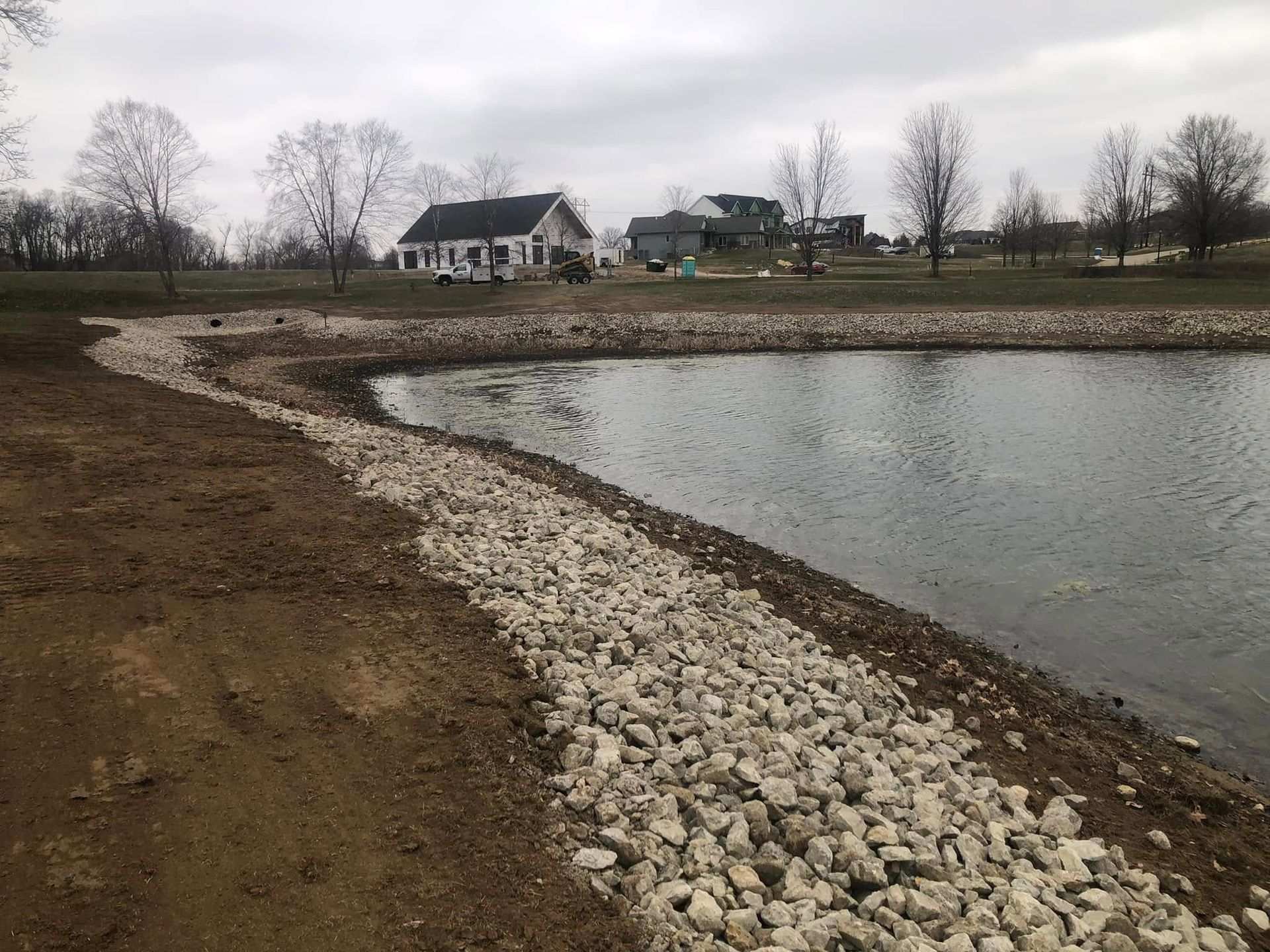 A large pond surrounded by rocks and dirt with a house in the background.