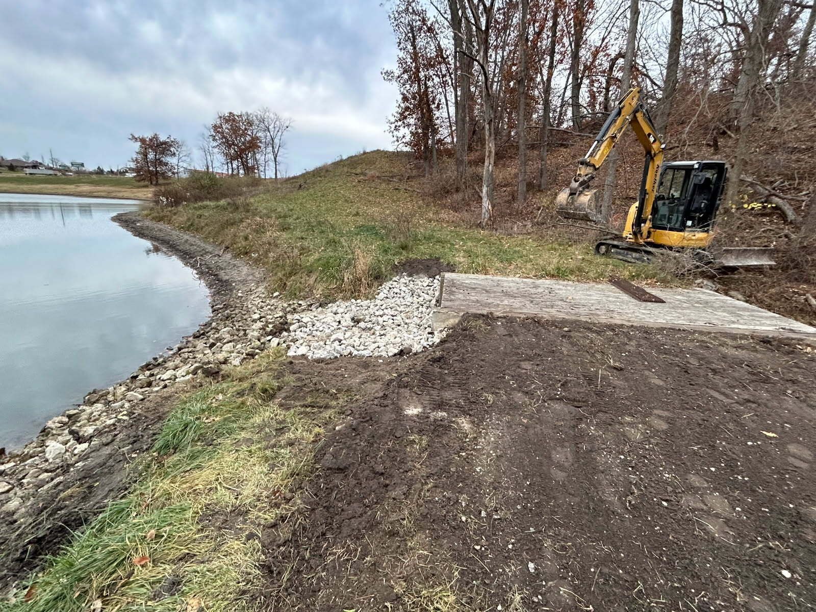 A small excavator works near a lake, with stone and dirt beside a paved area, trees in the background.