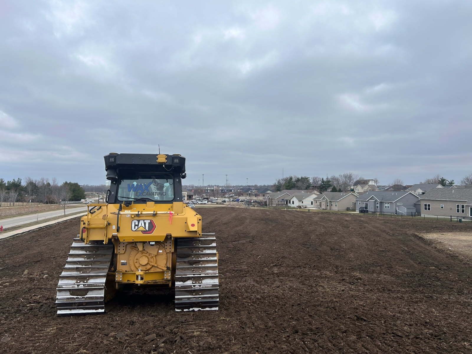 Yellow Caterpillar bulldozer on a brown soil mound, with houses and cloudy sky in the background.