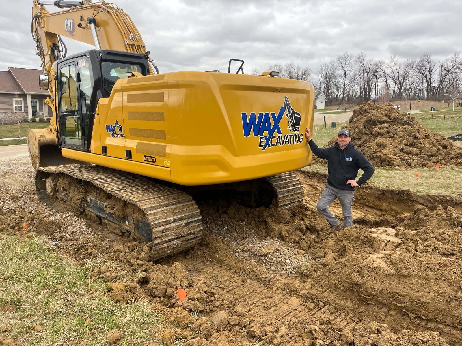 Man standing by yellow excavator digging a trench in a yard.