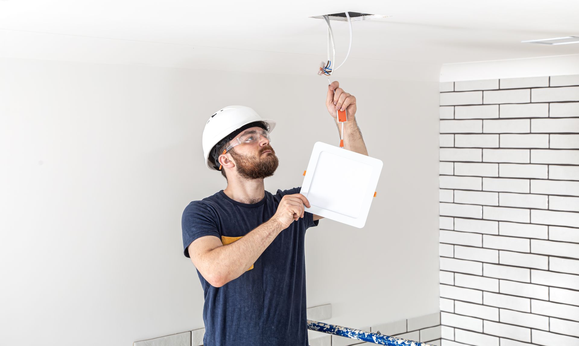 Electrician in hard hat, using screwdriver on ceiling wiring, holding plans.