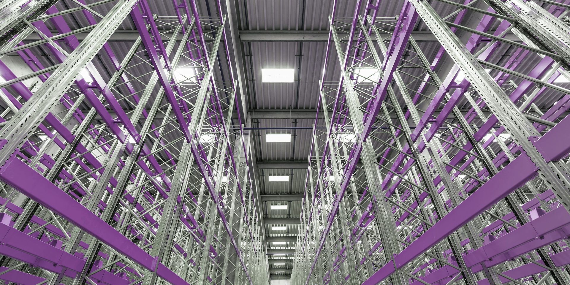 Warehouse interior with tall metal storage racks, a gray ceiling, and overhead lights. Purple accents.