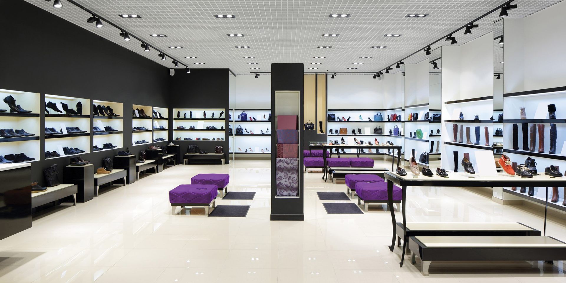 Shoe store interior. Shoes on shelves and display tables. Purple seating on the floor. Bright white and black accents.