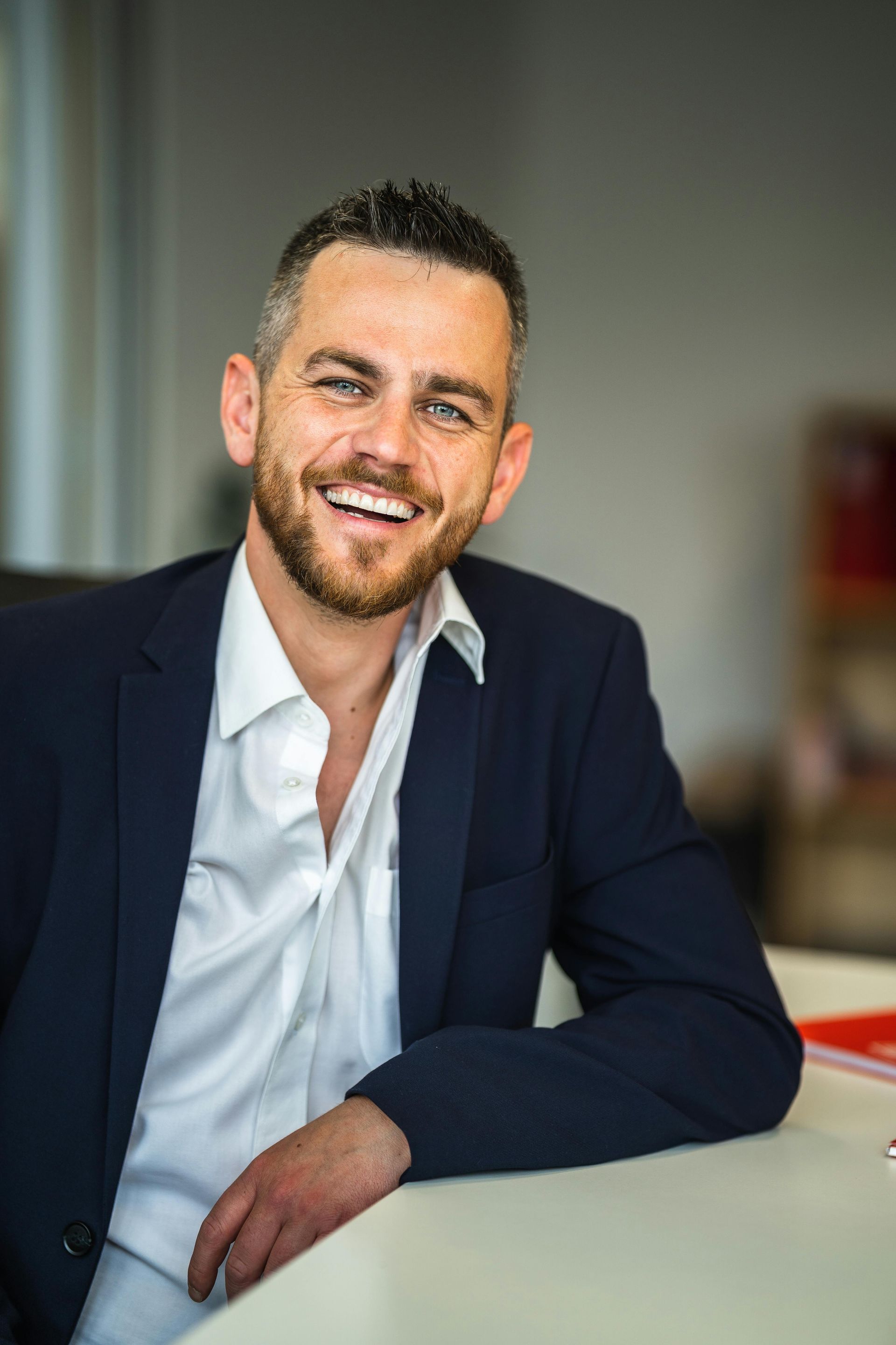 Man in a navy blazer and white shirt smiles widely at the camera, indoors.