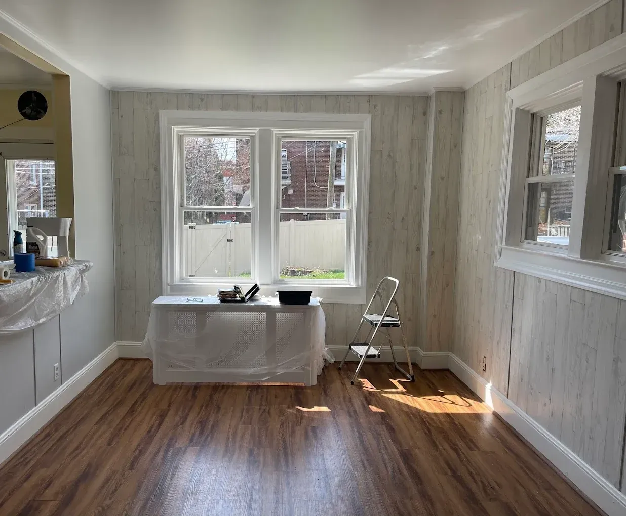 Interior of a room with wood floors, two windows, and textured wall paneling; a small step ladder and paint supplies are present.
