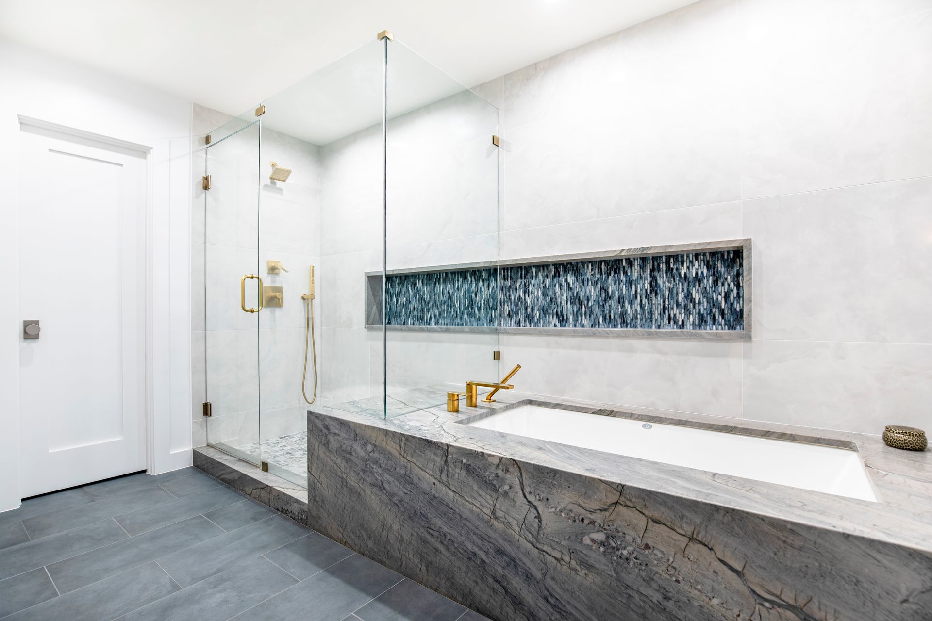 Modern bathroom with gray stone tub and shower, glass doors, and blue tile accent.