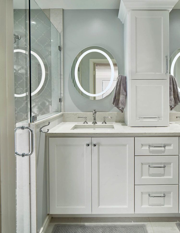 Bathroom with white vanity, tall cabinet, and oval mirror with illuminated frame.