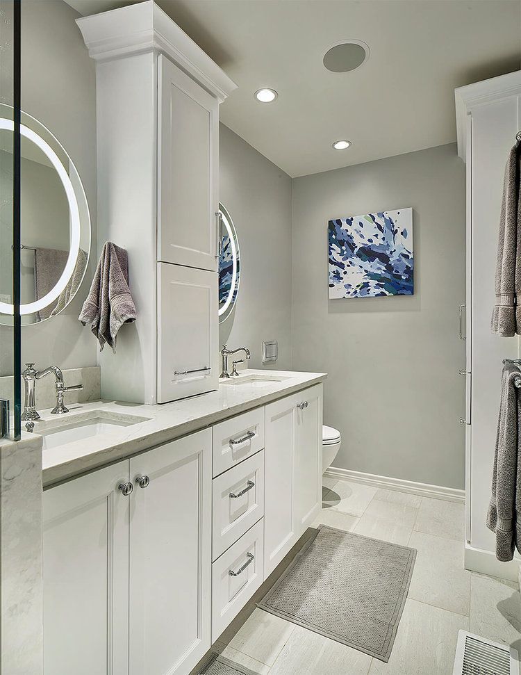 White bathroom with double vanity, tall storage cabinet, grey walls, art, and rug.