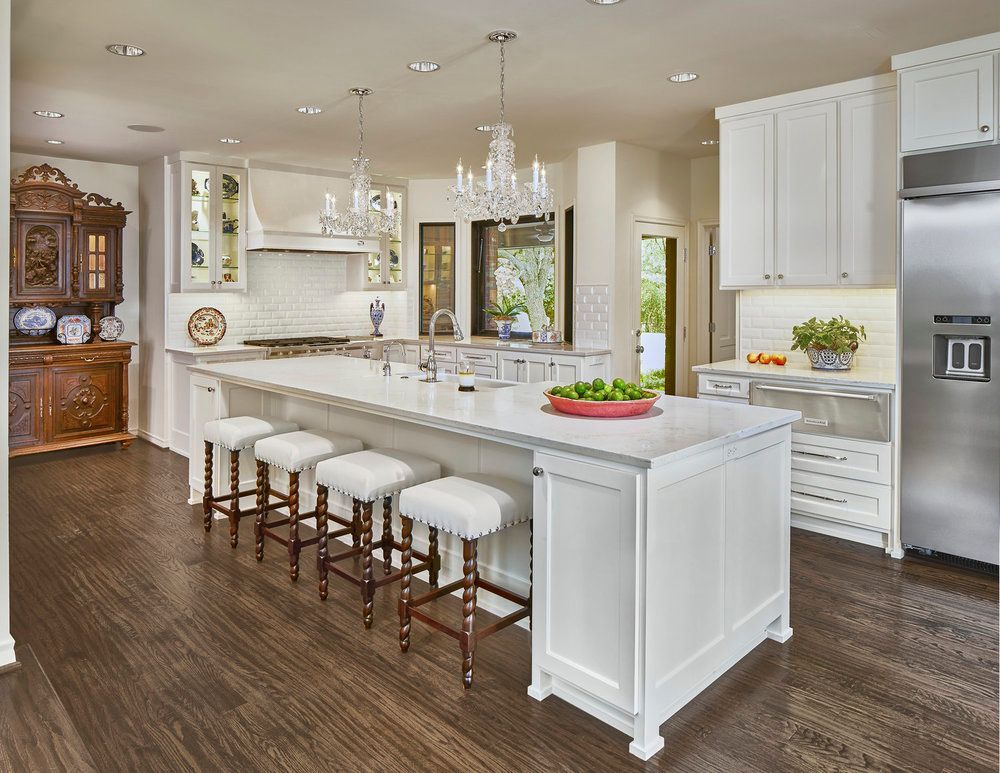 Bright white kitchen with large island, wood floors, crystal chandeliers, and stainless steel refrigerator.