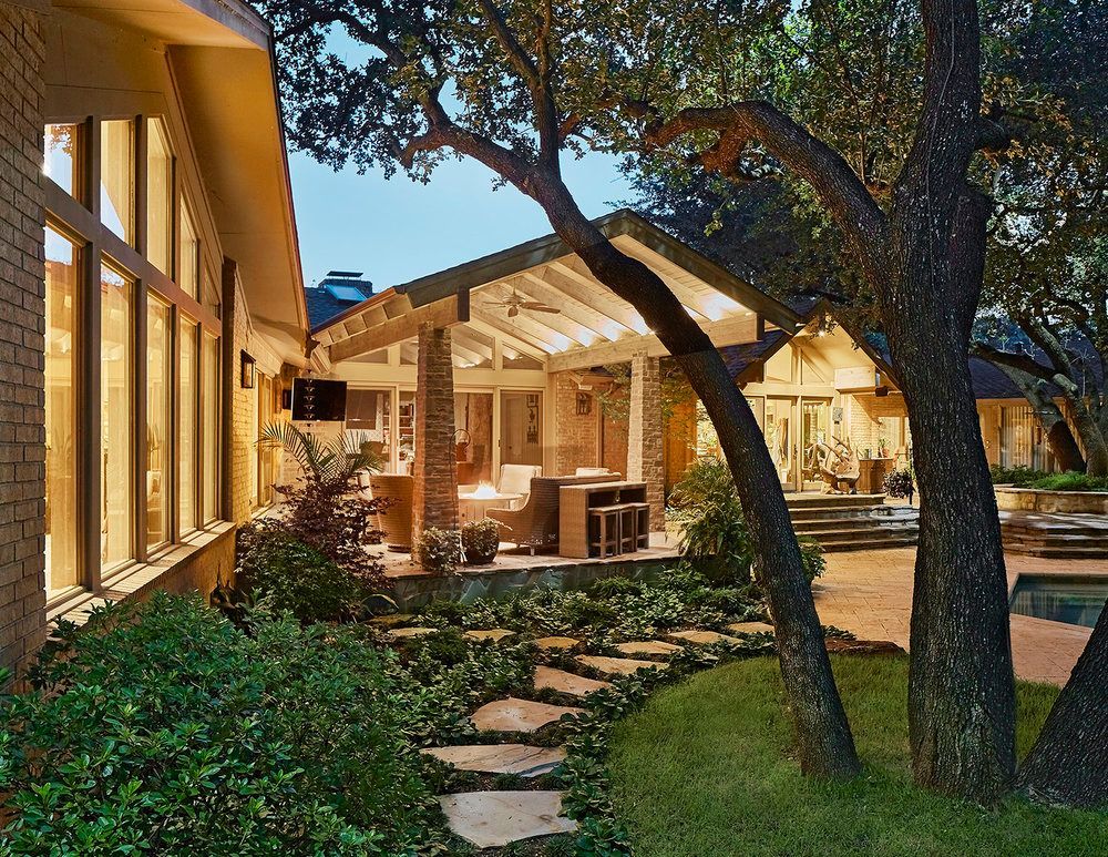 Backyard patio with stone walkway, green lawn, trees, and illuminated house at dusk.