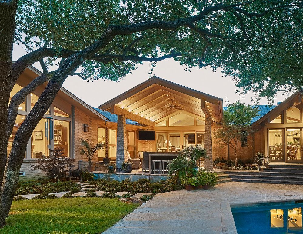 Backyard with covered patio, pool, and house with large windows. Evening light, lush landscaping.