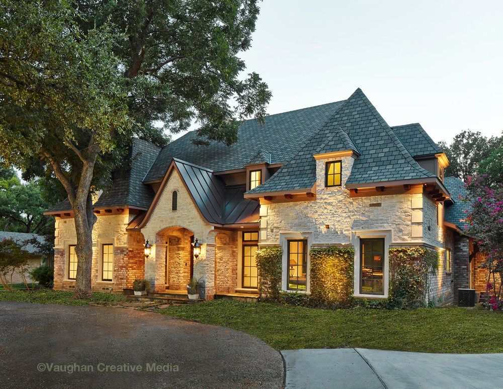 Stone and dark roofed house with a curving driveway, surrounded by greenery.