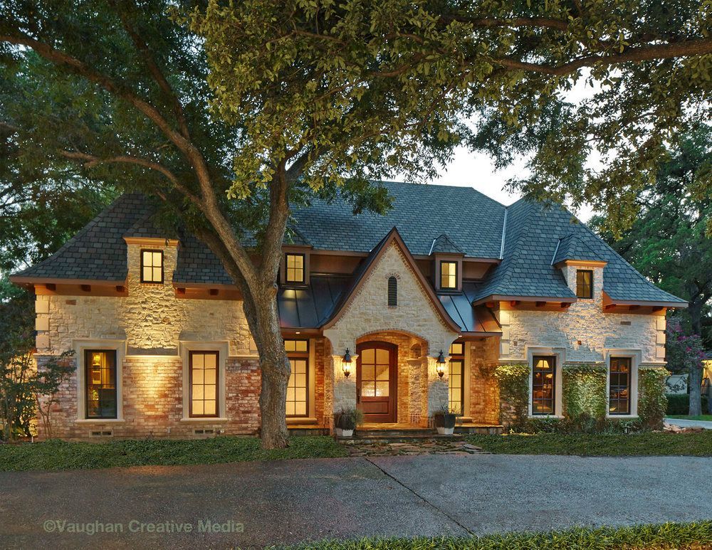 Stone house with illuminated windows and landscaping; a large tree in front.