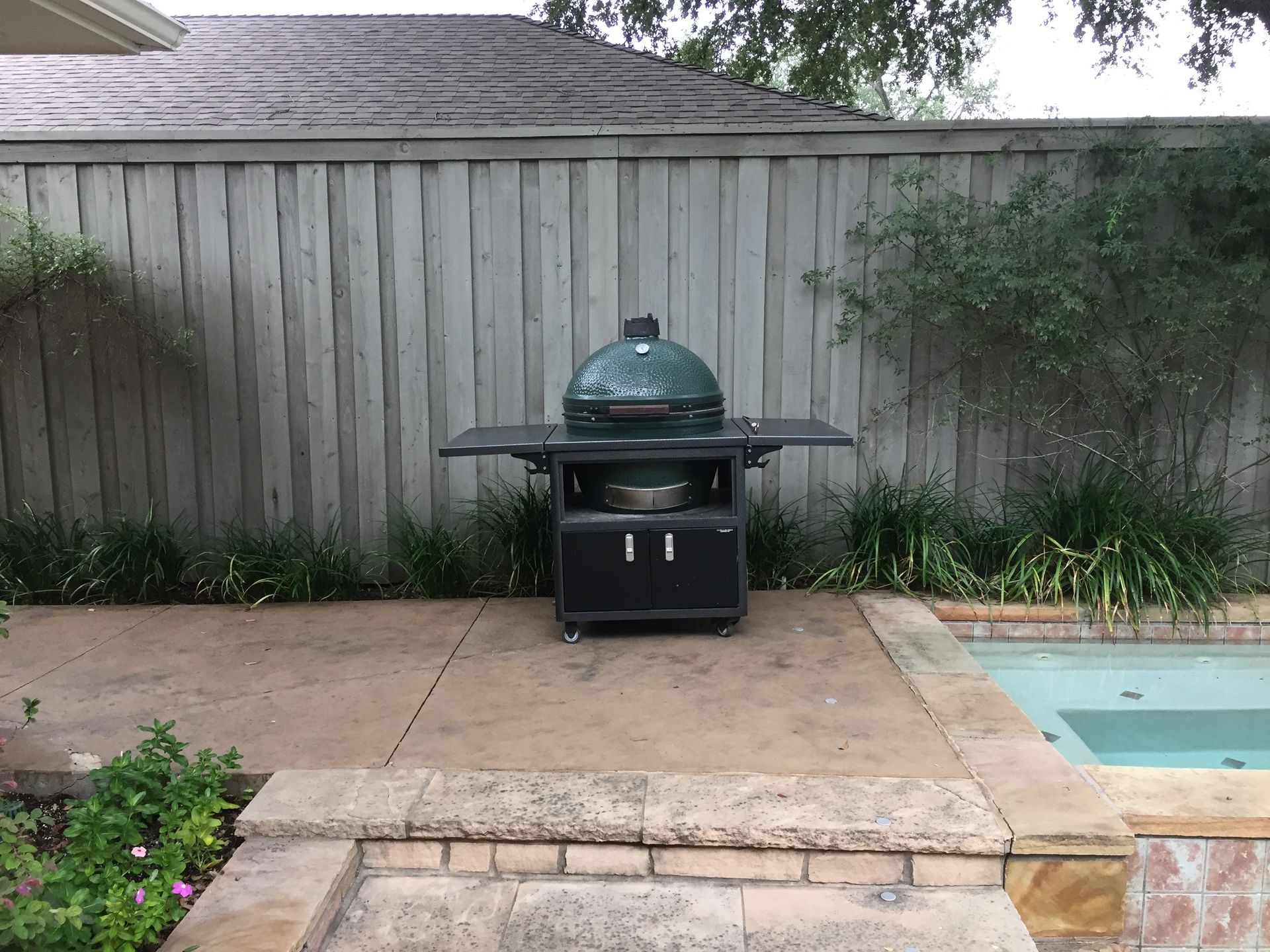 A green kamado-style grill sits on a black rolling cart on a backyard patio next to a pool.