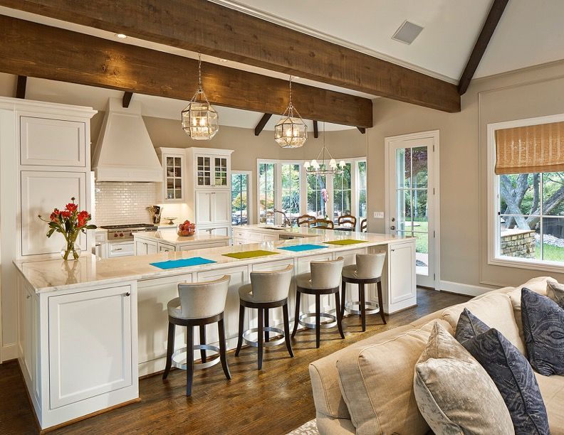Elegant white kitchen with island seating, pendant lights, wooden beams, and a view of a backyard.