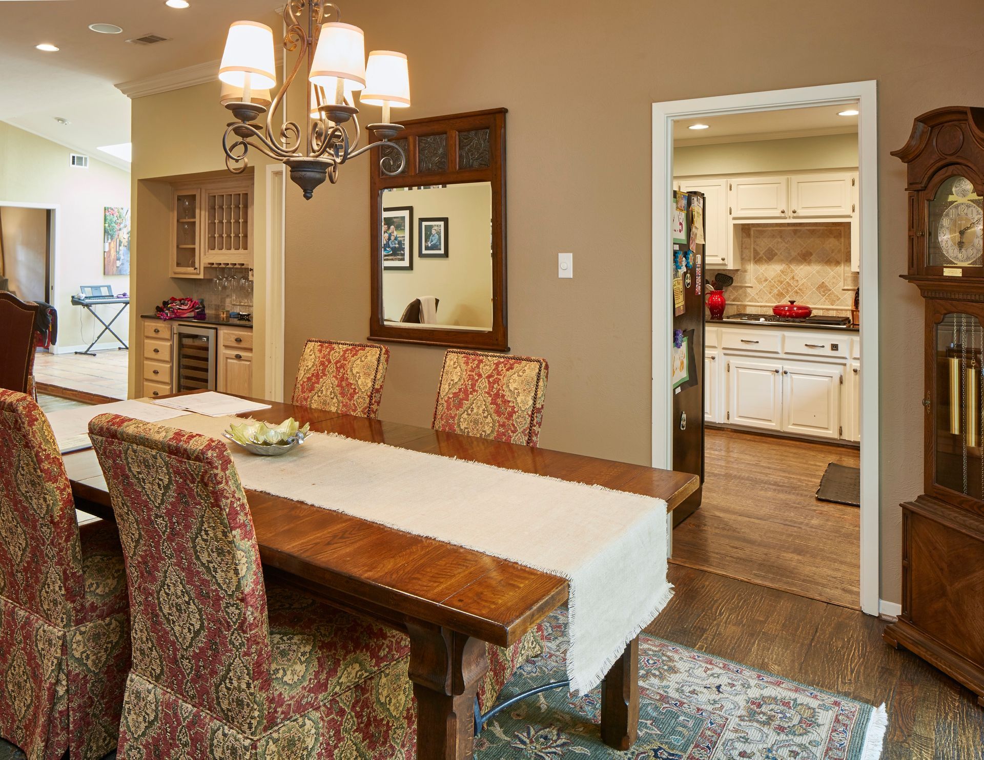 A dining room with a wooden table, patterned chairs, a chandelier, a wall mirror, and a grandfather clock.