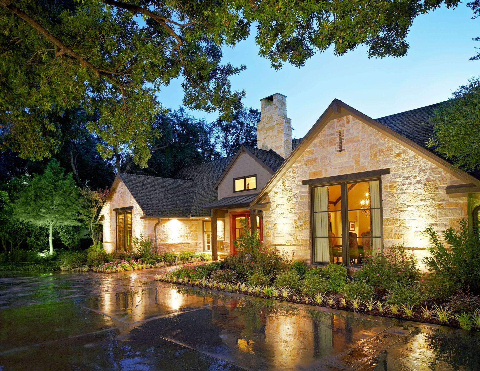 Stone house with lit exterior, wet driveway, and lush greenery. University Park exterior renovation.