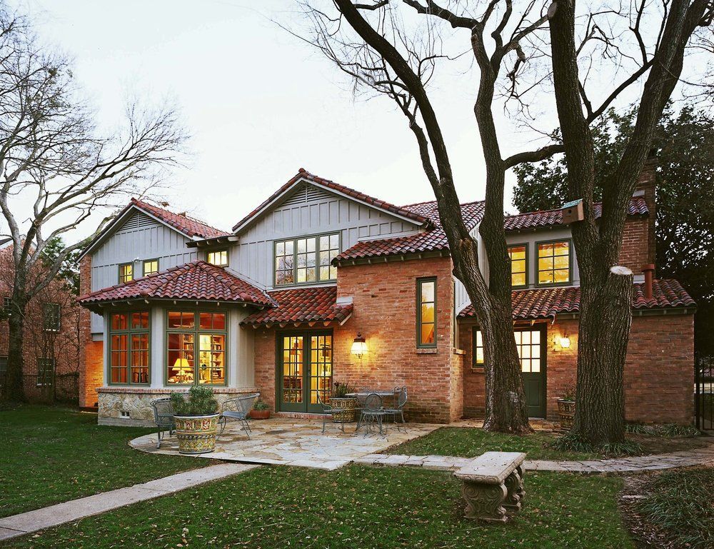 Red-brick house with terracotta roof, windows, and a stone patio. A large tree stands in the yard.