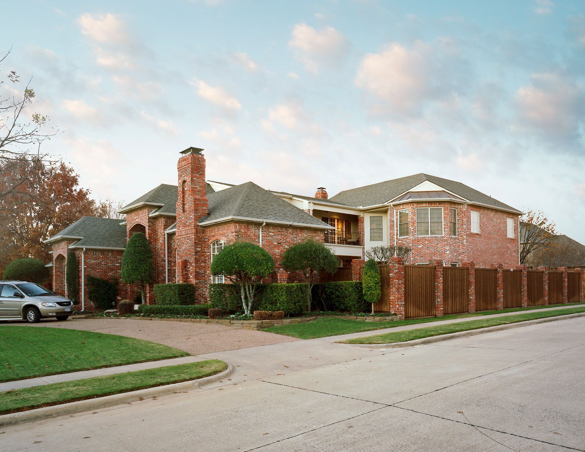 Large brick house with attached garage, green lawn, and brown fence under a blue sky.