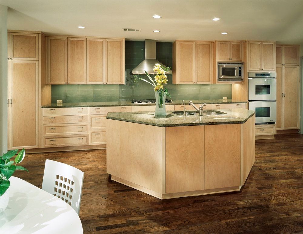 Modern kitchen with light wood cabinets, granite island, stainless steel appliances, and dark wood floors.