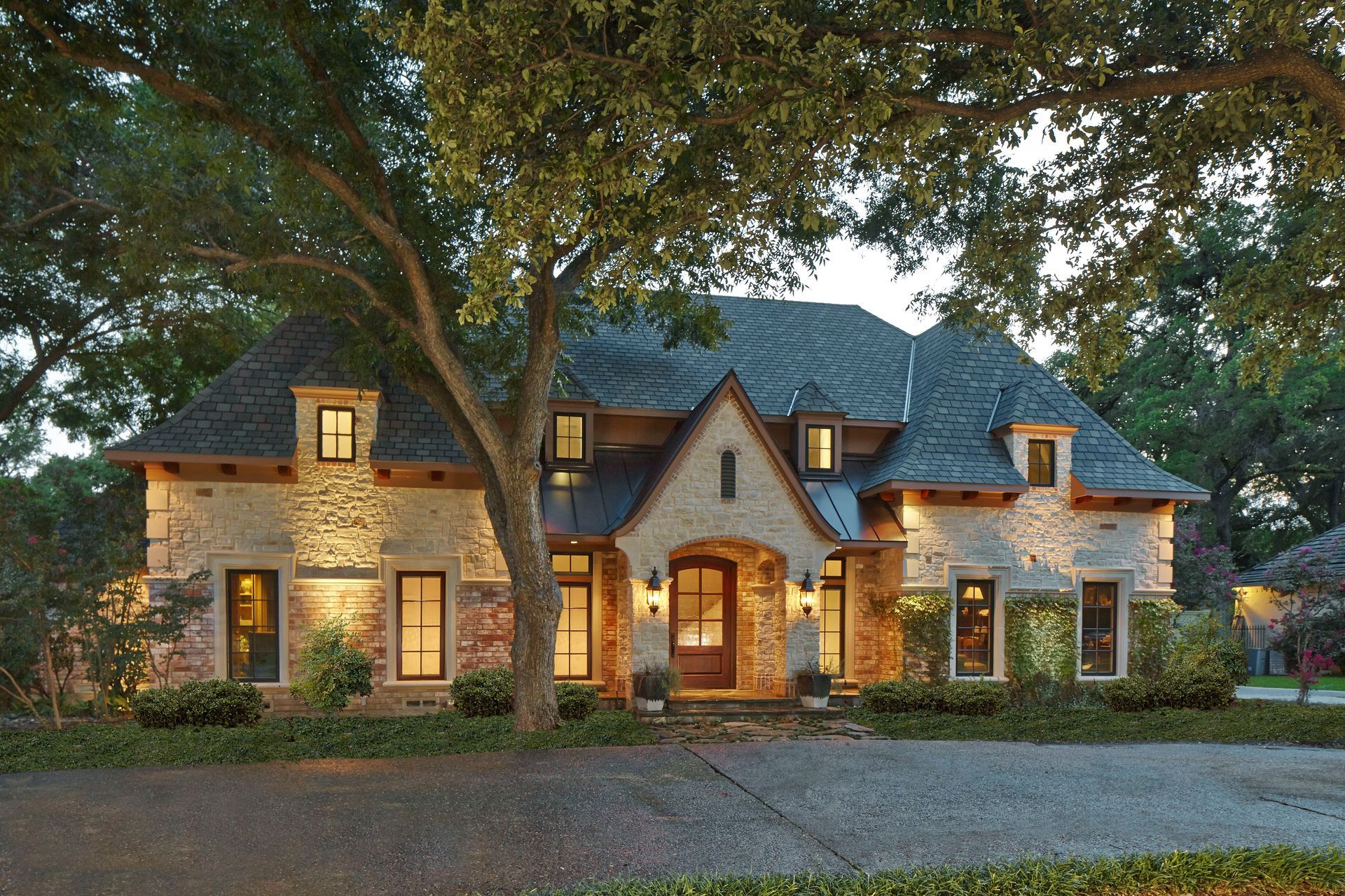Stone-faced house with dormers, lit warmly at dusk, fronted by a driveway and large tree.