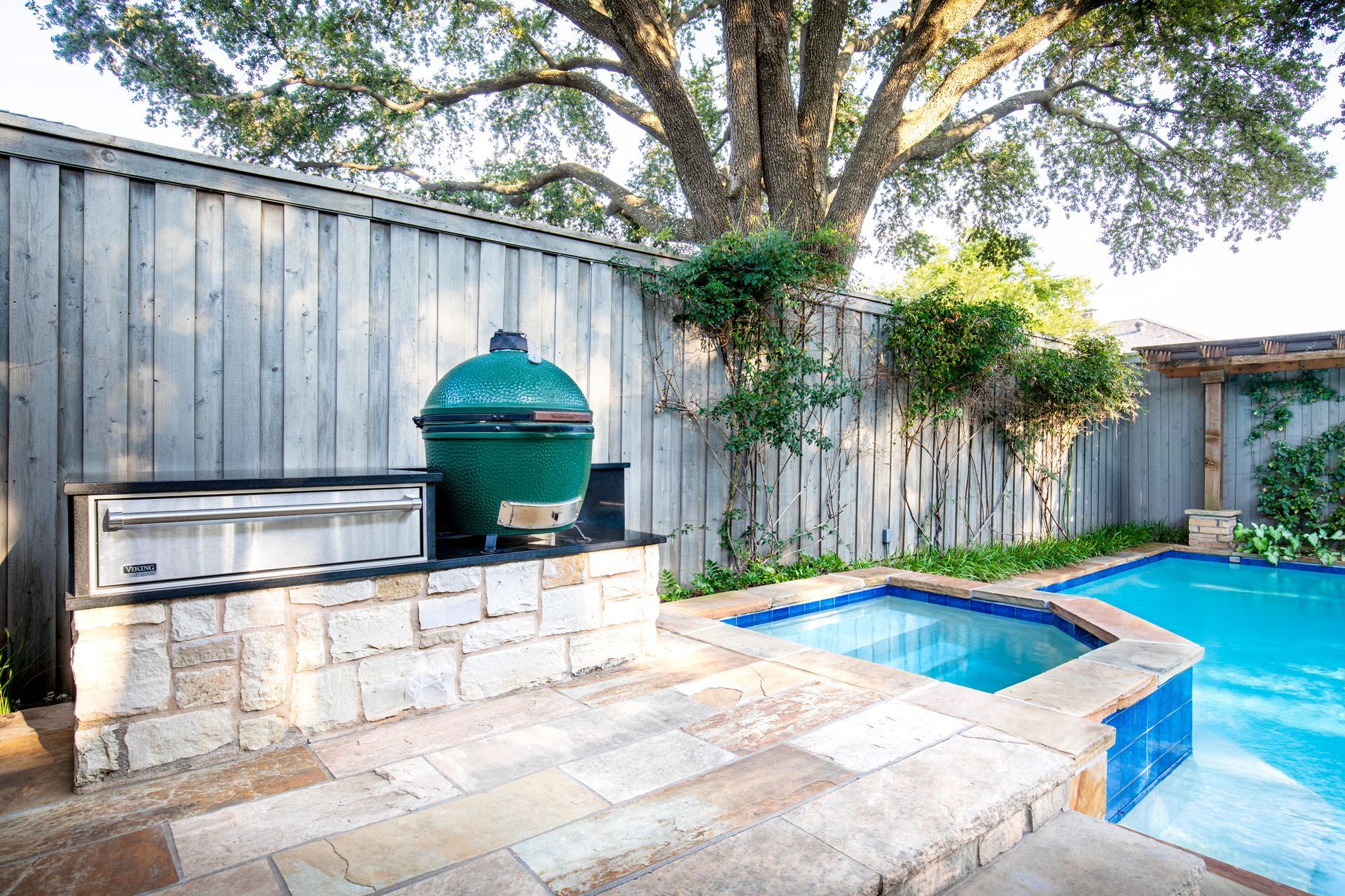 Outdoor kitchen with a green ceramic grill and stainless steel drawers next to a backyard pool and hot tub.