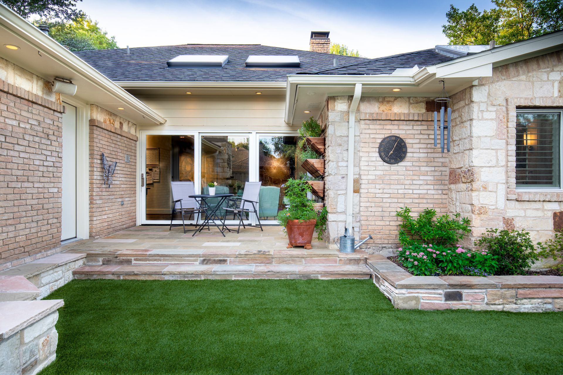 A stone house with a backyard featuring a patio, outdoor seating, a small patch of artificial turf, and potted plants.
