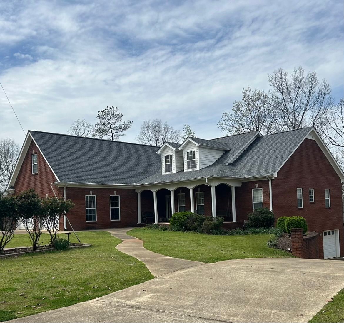 Brick house with dark roof and white columns, surrounded by green grass and a concrete driveway.