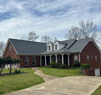 Roofer kneels, installing asphalt shingles on a house roof. He wears a baseball cap and uses a pneumatic nail gun.