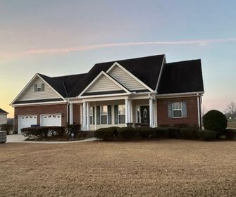 A one-story brick house with a dark roof and a front porch. Two-car garage, manicured lawn. Evening sky.