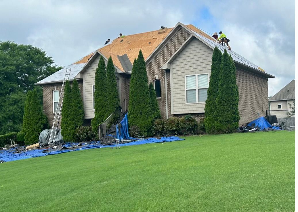 House with roofing construction in progress; workers on roof, blue tarps protect lawn.